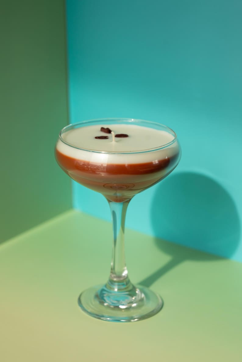 K&ouml;nnte beinhalten: Ein klares Cocktailglas gef&uuml;llt mit einem geschichteten Getr&auml;nk. Die untere Schicht ist dunkelbraun, bedeckt mit einer wei&szlig;en Schicht und garniert mit drei dunkelbraunen Kaffeebohnen. Das Glas steht auf einer gr&uuml;nen und blauen Oberfl&auml;che.