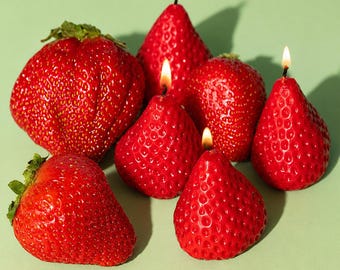 May include: A collection of red strawberry-shaped candles with lit wicks, arranged on a light green surface. The candles vary in size, with some resembling actual strawberries. The image captures the candles' texture and vibrant color.