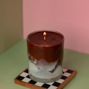 May include: A lit candle in a clear glass with a layered design of brown and white. The candle sits on a square coaster with a black and white checkered pattern. The background is a mix of pink and green.