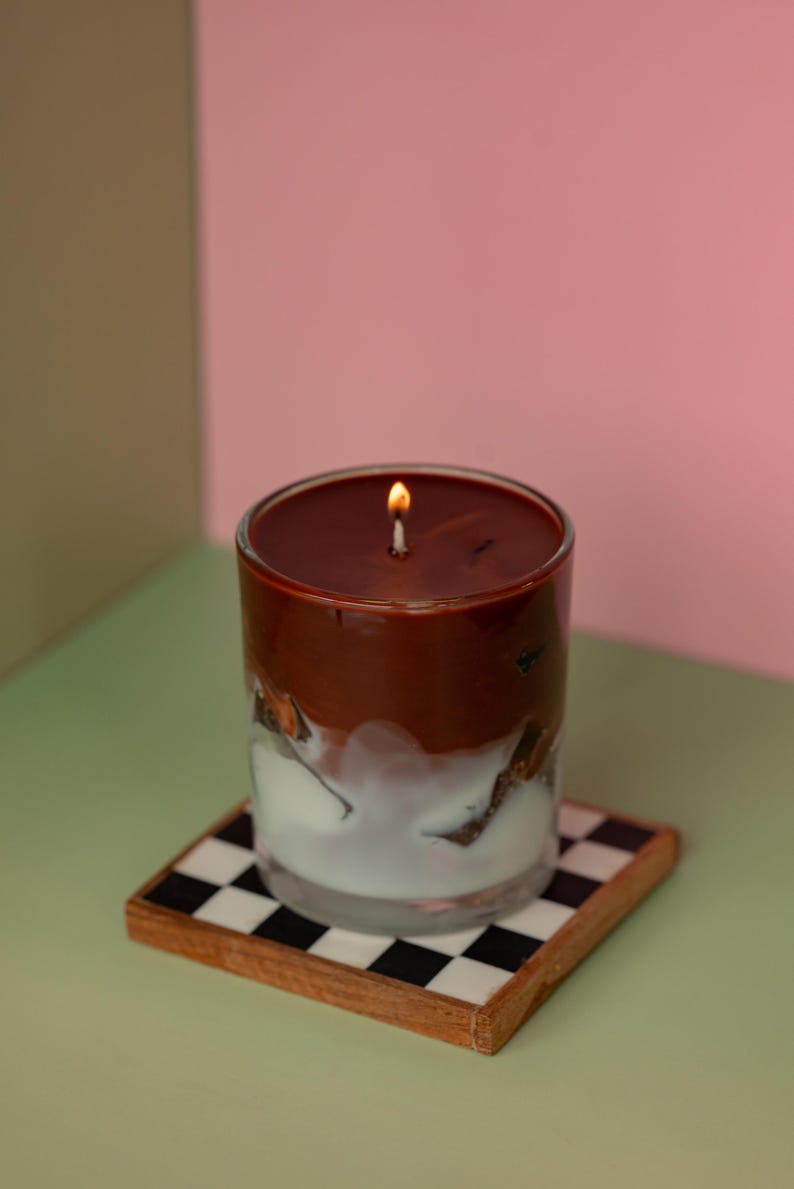 May include: A lit candle in a clear glass with a layered design of brown and white. The candle sits on a square coaster with a black and white checkered pattern. The background is a mix of pink and green.