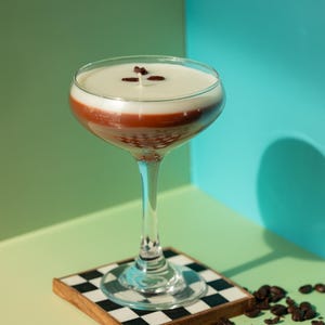 K&ouml;nnte beinhalten: Ein geschichteter Cocktail in einem Stielglas, mit einer wei&szlig;en oberen Schicht, einer rotbraunen Mitte und einer klaren Basis. Das Getr&auml;nk ist mit Kaffeebohnen garniert und steht auf einem karierten Untersetzer. Kaffeebohnen sind verstreut.