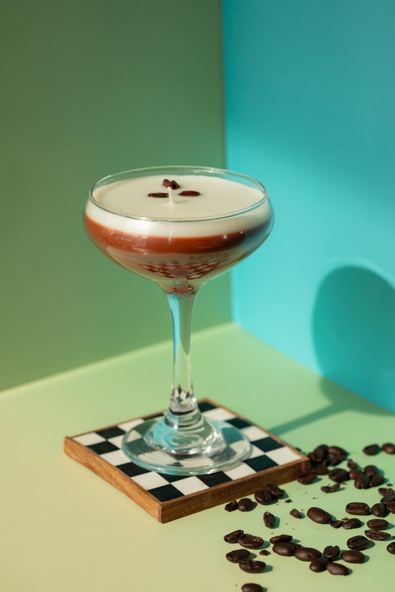 K&ouml;nnte beinhalten: Ein geschichteter Cocktail in einem Stielglas, mit einer wei&szlig;en oberen Schicht, einer rotbraunen Mitte und einer klaren Basis. Das Getr&auml;nk ist mit Kaffeebohnen garniert und steht auf einem karierten Untersetzer. Kaffeebohnen sind verstreut.