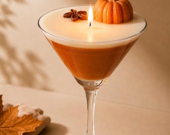 May include: A glass martini-style candle featuring a two-toned wax design with a dark orange base and a creamy white top layer. The candle is lit and topped with a small orange pumpkin-shaped wax accent and a single brown star anise spice.