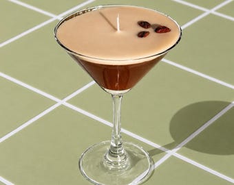 May include: A candle in the shape of a martini glass, featuring a brown wax base and a tan top. It has a white wick and is adorned with three dark brown coffee beans. The glass is placed on a green tiled surface.