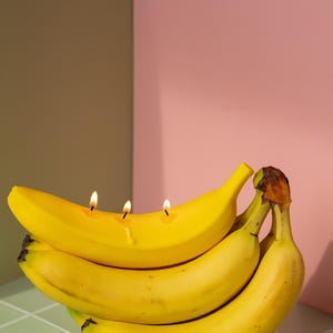 Pode incluir: Um cacho de bananas amarelas maduras, com três velas acesas inseridas em uma delas. As bananas estão sobre uma superfície de azulejos verdes e brancos, com um fundo rosa e marrom.