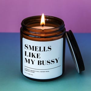 May include: A brown glass candle jar with a white label that reads "Smells Like My Bussy". The candle is lit and the jar has a black lid.