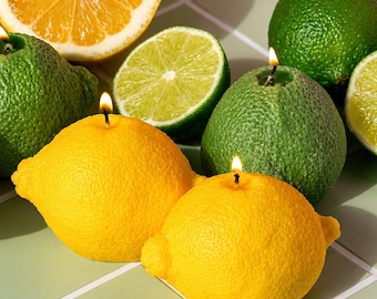 Puede incluir: Una naturaleza muerta con velas con forma de fruta y cítricos frescos. Dos velas amarillas con forma de limón con mechas encendidas en primer plano. Velas verdes con forma de lima y cítricos cortados por la mitad, incluyendo naranjas y limas, están dispuestas sobre una superficie embaldosada.