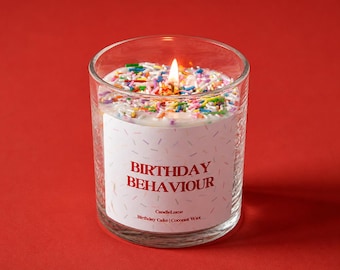 以下が含まれることがあります： 赤い背景に、"BIRTHDAY BEHAVIOUR"と赤い文字で書かれた白いラベルが付いた、透明なガラス瓶に入った点灯したキャンドル。キャンドルのワックスはカラフルなスプリンクルで覆われています。