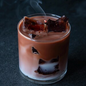 May include: A candle in a clear glass, designed to resemble a cocktail. The candle has a layered appearance with a white base, a light brown middle, and a dark brown top. Dark brown wax cubes are scattered throughout, and smoke rises from the lit wick.
