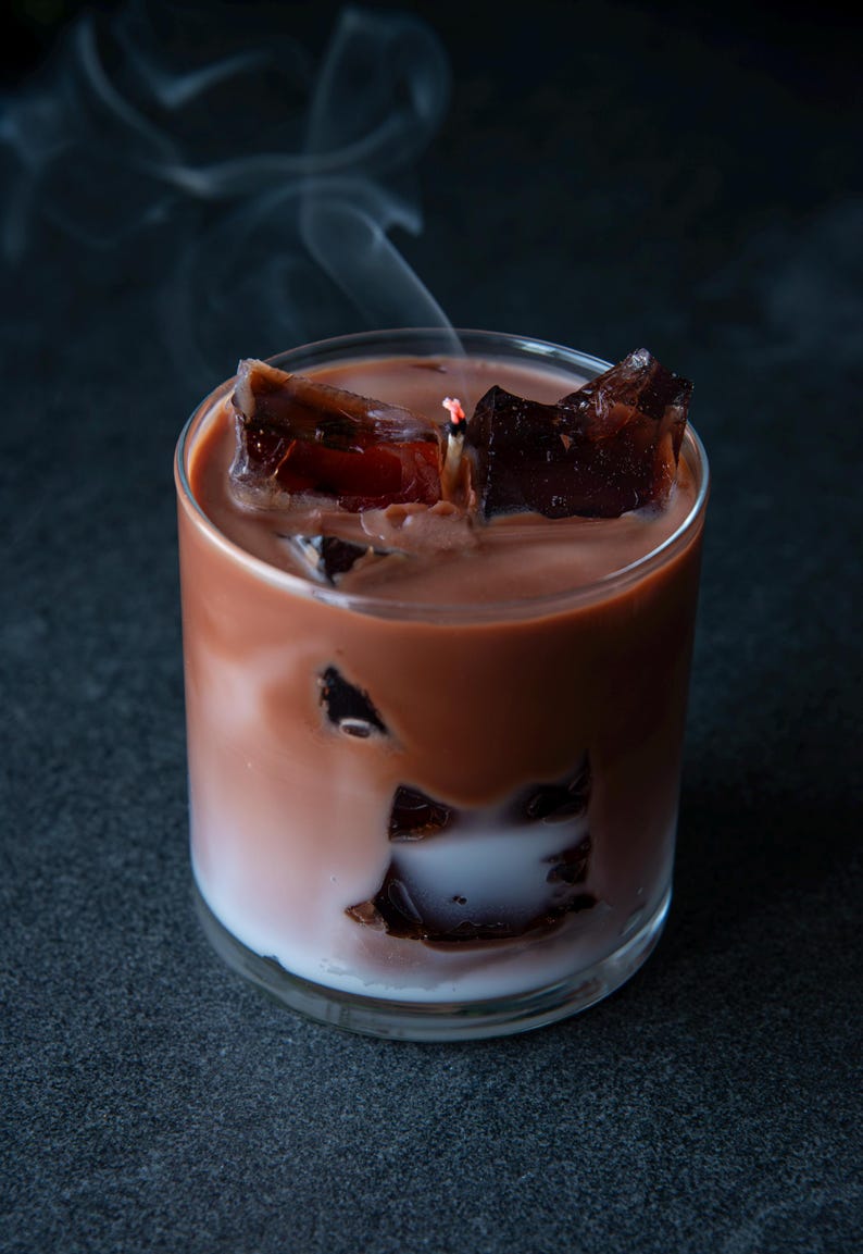 May include: A candle in a clear glass, designed to resemble a cocktail. The candle has a layered appearance with a white base, a light brown middle, and a dark brown top. Dark brown wax cubes are scattered throughout, and smoke rises from the lit wick.