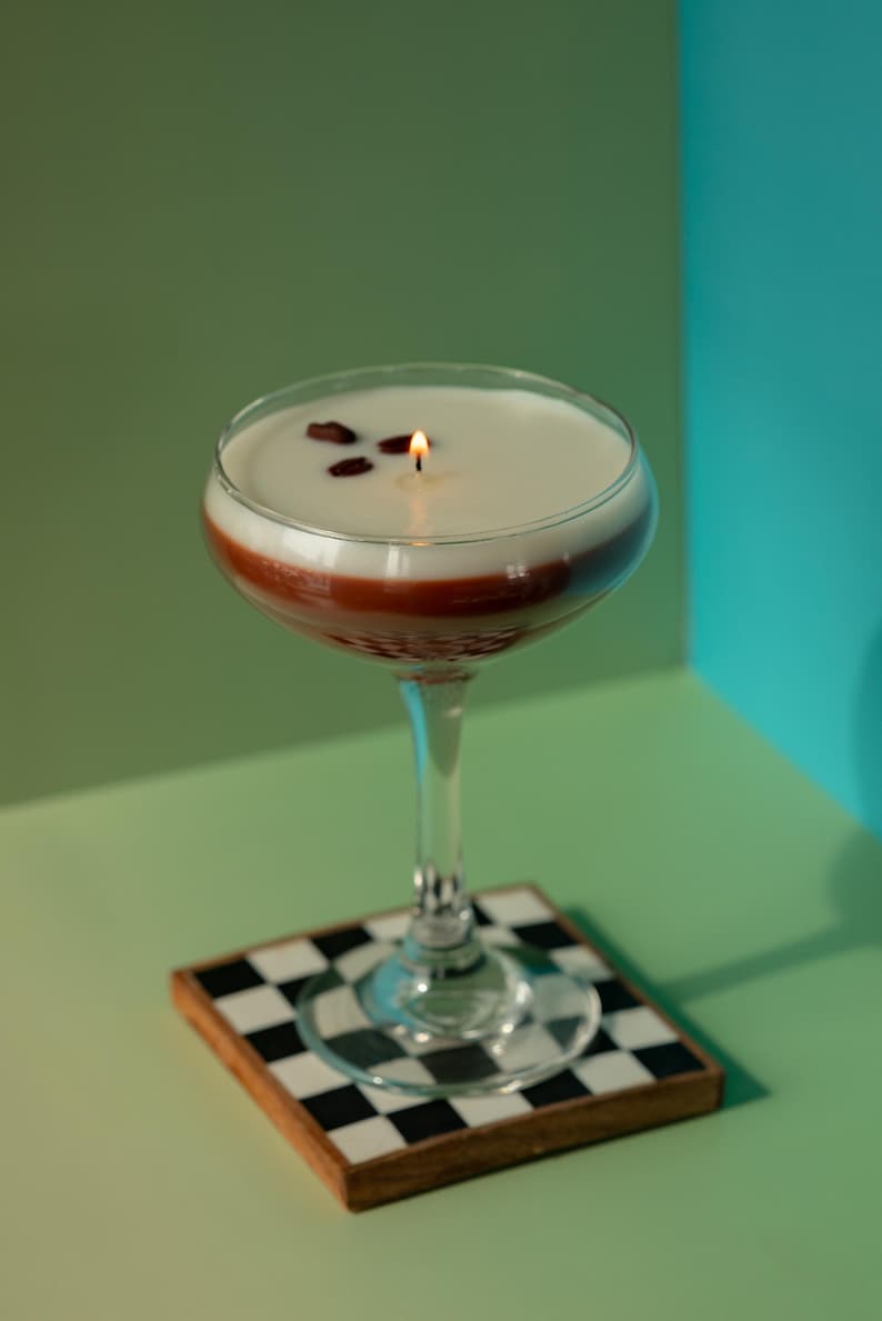 K&ouml;nnte beinhalten: Eine Kerze in Cocktailform mit einer wei&szlig;en oberen Schicht, einer roten Mittelschicht und einem transparenten Stiel. Die Kerze ist angez&uuml;ndet, mit drei Kaffeebohnen obenauf. Sie steht auf einem karierten Untersetzer.