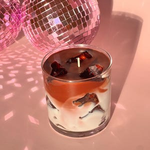 May include: A layered candle in a clear glass. The candle has a white base, a reddish-orange middle layer, and a dark brown top with embedded decorative pieces. A single wick is centered. Two pink disco balls are in the background.