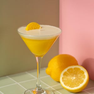 May include: A yellow cocktail in a martini glass, garnished with a lemon slice, sits on a tiled surface. Two lemons, one halved, are next to the glass. The background features a pink and olive-green wall.
