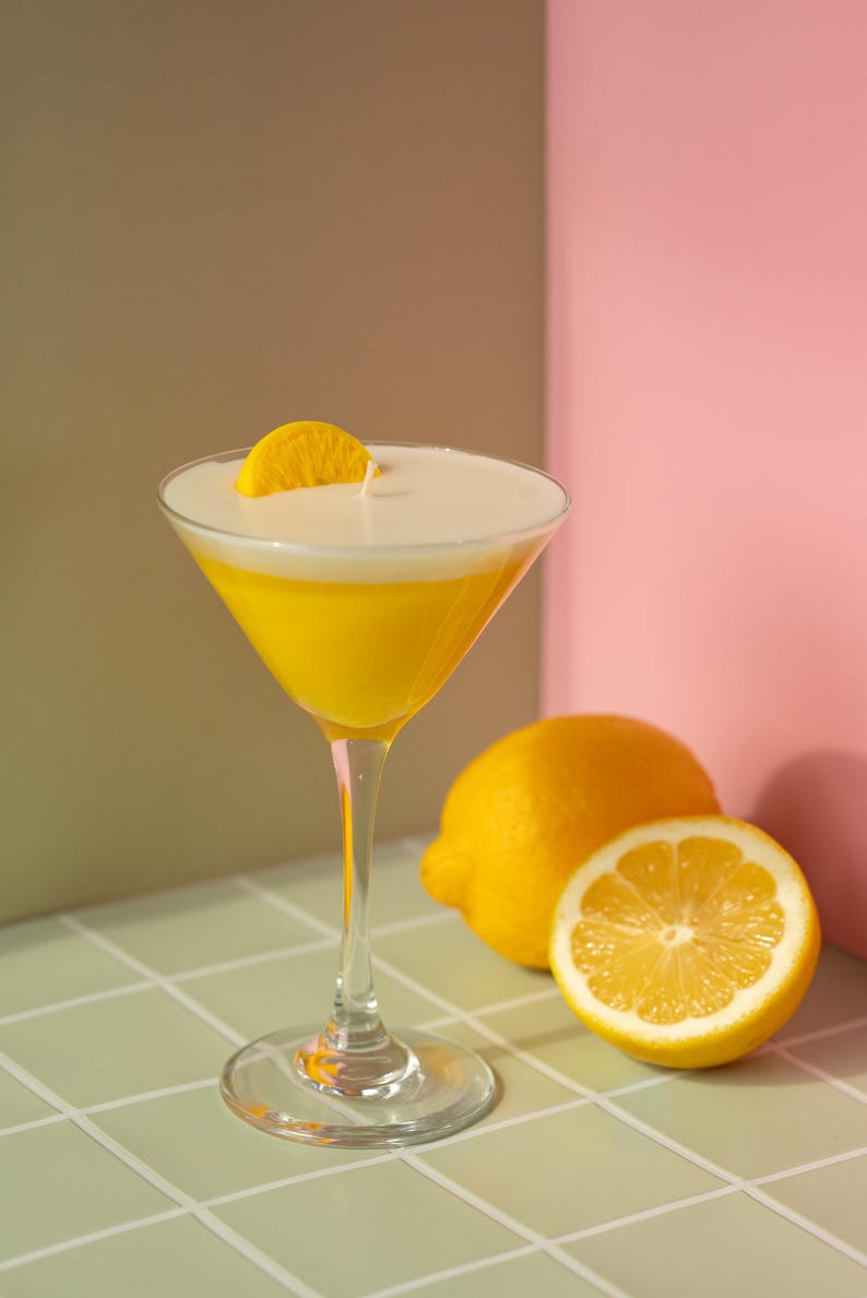 May include: A yellow cocktail in a martini glass, garnished with a lemon slice, sits on a tiled surface. Two lemons, one halved, are next to the glass. The background features a pink and olive-green wall.