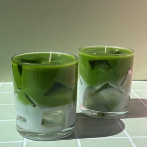 May include: Two clear glass candles with a layered design. The top layer is a vibrant green, while the bottom layer is white, creating a visually appealing contrast. The candles have a smooth, glossy finish and a simple, elegant aesthetic.
