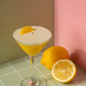 May include: A yellow cocktail candle in a martini glass, garnished with a lemon slice. The candle has a white top and a lit wick. Two lemons, one halved, sit nearby on a tiled surface. The background is a mix of green, brown, and pink.