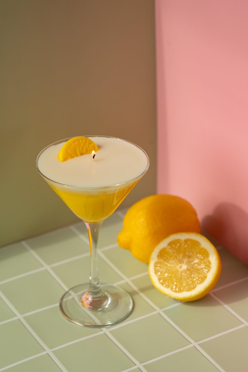May include: A yellow cocktail candle in a martini glass, garnished with a lemon slice. The candle has a white top and a lit wick. Two lemons, one halved, sit nearby on a tiled surface. The background is a mix of green, brown, and pink.