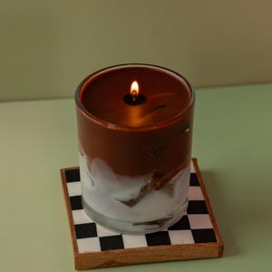 May include: A lit candle in a clear glass jar with a brown top and white bottom. The candle sits on a small square coaster with a black and white checkerboard pattern. The background is a solid light green.