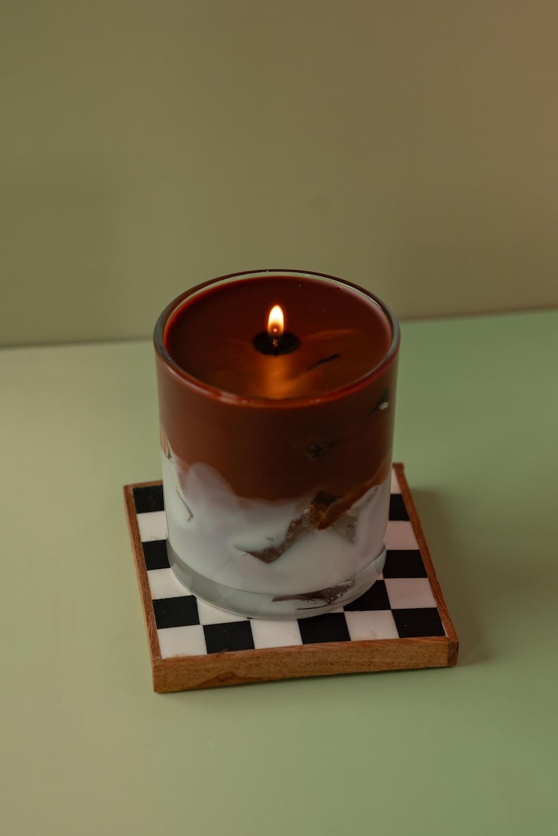 May include: A lit candle in a clear glass jar with a brown top and white bottom. The candle sits on a small square coaster with a black and white checkerboard pattern. The background is a solid light green.