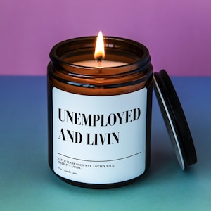 May include: A brown glass candle jar with a white label that reads "Unemployed and Livin". The candle is lit and the jar has a black lid.