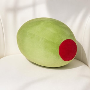 Puede incluir: Un cojín de felpa con forma de aceituna de gran tamaño. El cojín es principalmente de color verde claro con un gran acento rojo redondo en un extremo. Descansa sobre una superficie blanca, lo que sugiere un artículo decorativo o novedoso.