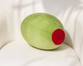 May include: A plush, oversized olive-shaped pillow. The pillow is primarily light green with a large, round, red accent on one end. It rests on a white surface, suggesting a decorative or novelty item.