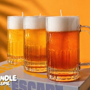May include: Three beer-shaped candles with white foam tops and amber-colored wax. The candles are in clear glass mugs with handles, sitting on a book with the text "CANDLE LUME". The background is a solid orange.