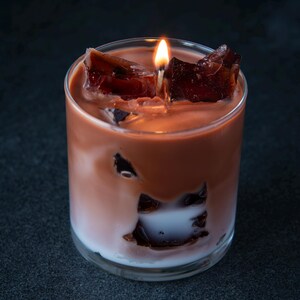 May include: A candle in a clear glass, resembling a cocktail. The candle has a layered appearance with a white base, a light brown middle, and dark brown chunks on top. A lit wick sits in the center.