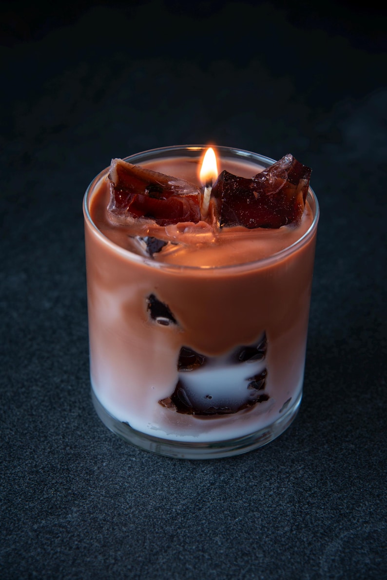May include: A candle in a clear glass, resembling a cocktail. The candle has a layered appearance with a white base, a light brown middle, and dark brown chunks on top. A lit wick sits in the center.