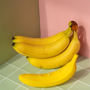 以下が含まれることがあります： 熟した黄色のバナナの房で、手前に1本のバナナがあります。バナナは、ピンクと茶色の背景に、薄緑色のタイルの上にあります。バナナは鮮やかな黄色です。