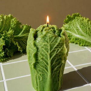 Puede incluir: Una vela de cera verde con forma de lechuga, con hojas detalladas y texturizadas y una mecha encendida en la parte superior. La vela está colocada en posición vertical, mostrando una forma vegetal realista con venas y bordes rizados.