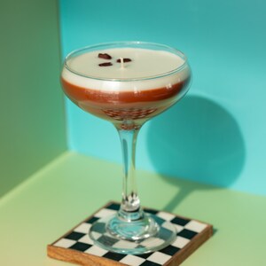 K&ouml;nnte beinhalten: Eine Cocktail-Kerze in einem Coup&eacute;-Glas, mit einem geschichteten Design aus Wei&szlig;, Braun und klarem Glas. Sie steht auf einem karierten Untersetzer mit Holzrahmen. Der Hintergrund ist ein Farbverlauf aus Gr&uuml;n und Blau.