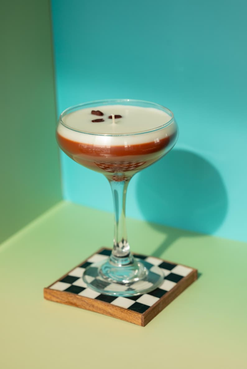 K&ouml;nnte beinhalten: Eine Cocktail-Kerze in einem Coup&eacute;-Glas, mit einem geschichteten Design aus Wei&szlig;, Braun und klarem Glas. Sie steht auf einem karierten Untersetzer mit Holzrahmen. Der Hintergrund ist ein Farbverlauf aus Gr&uuml;n und Blau.