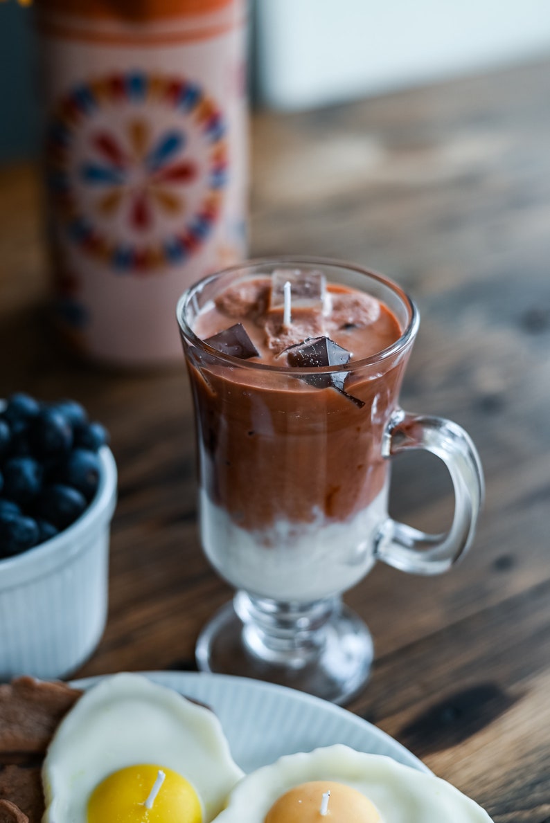 May include: A glass mug filled with a brown and white layered candle, resembling a coffee drink with ice cubes. The candle has a white wick. The mug is on a wooden table next to a white plate with two fried egg candles.