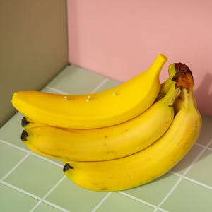 以下が含まれることがあります： 茶色の先端が付いた熟した黄色のバナナの束が、明るい緑色のタイルの表面に配置されています。バナナはピンクとオリーブグリーンの背景の前にあります。バナナには小さな白いキャンドルの芯が付いています。