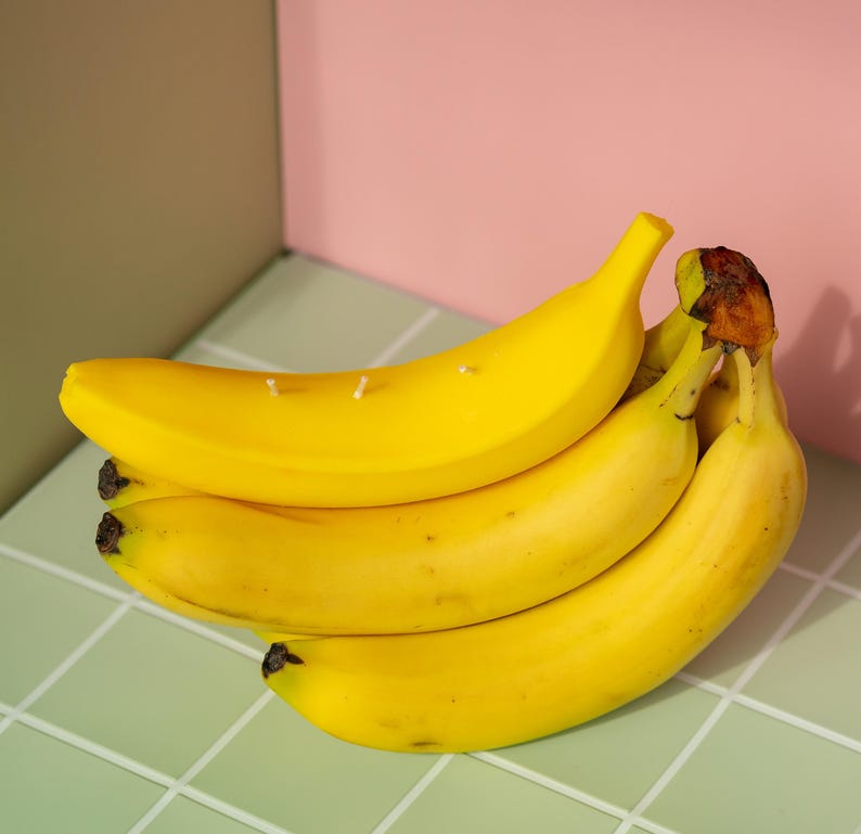 以下が含まれることがあります： 茶色の先端が付いた熟した黄色のバナナの束が、明るい緑色のタイルの表面に配置されています。バナナはピンクとオリーブグリーンの背景の前にあります。バナナには小さな白いキャンドルの芯が付いています。