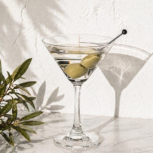 May include: A clear martini glass filled with a clear liquid and two green olives. A black cocktail pick with a small black ball on the end rests in the glass. The glass sits on a marble surface.