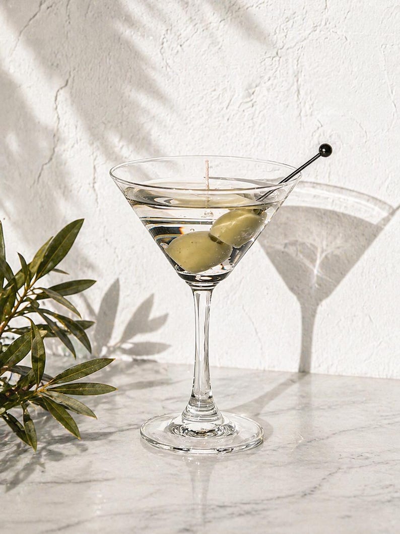 May include: A clear martini glass filled with a clear liquid and two green olives. A black cocktail pick with a small black ball on the end rests in the glass. The glass sits on a marble surface.