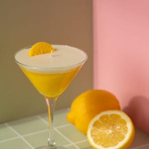 May include: A yellow cocktail in a martini glass, garnished with a lemon slice. The drink sits on a tiled surface next to a whole lemon and a lemon half. The background is a mix of green, brown, and pink.