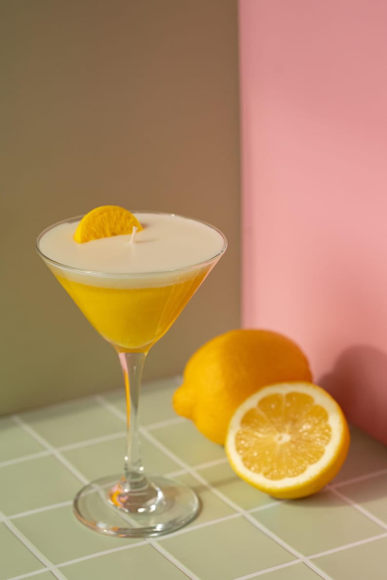 May include: A yellow cocktail in a martini glass, garnished with a lemon slice. The drink sits on a tiled surface next to a whole lemon and a lemon half. The background is a mix of green, brown, and pink.