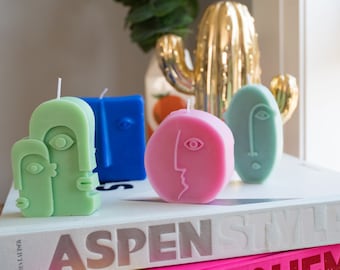 Peut inclure: Quatre bougies en forme de visage abstrait dans différentes couleurs et formes. Les bougies sont vertes, bleues, roses et bleu clair. Les bougies sont posées sur un livre blanc avec le texte "Aspen Style" imprimé sur la couverture.