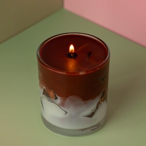 May include: A lit candle in a clear glass jar. The candle has a two-tone design with a dark brown top and a white, textured bottom. The flame glows brightly against the dark wax.