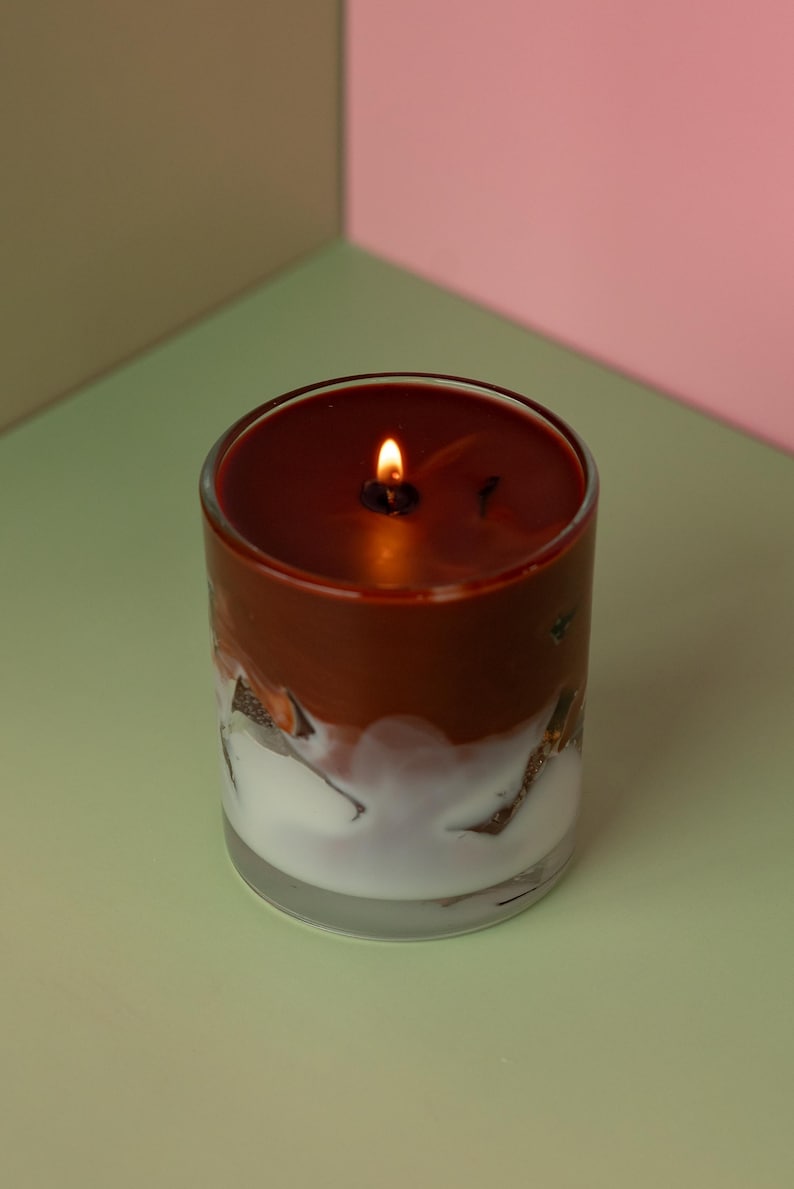 May include: A lit candle in a clear glass jar. The candle has a two-tone design with a dark brown top and a white, textured bottom. The flame glows brightly against the dark wax.