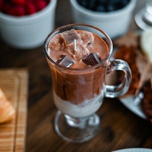 May include: A glass of iced chocolate with ice cubes and a straw. The drink is in a clear glass with a handle and is sitting on a wooden table.