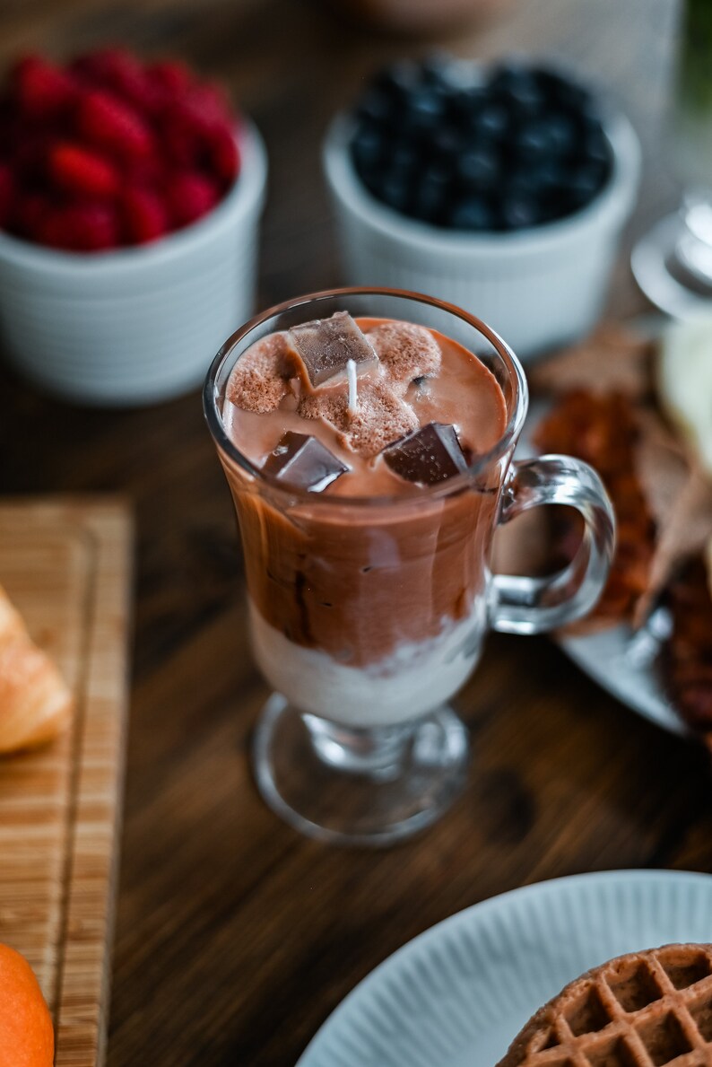 May include: A glass of iced chocolate with ice cubes and a straw. The drink is in a clear glass with a handle and is sitting on a wooden table.
