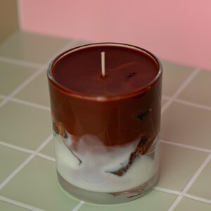 May include: A layered candle in a clear glass. The top layer is a dark brown, followed by a white layer, and a clear base. A white wick is centered on the top. The candle sits on a light green tiled surface.