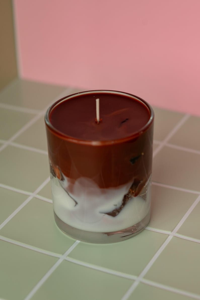May include: A layered candle in a clear glass. The top layer is a dark brown, followed by a white layer, and a clear base. A white wick is centered on the top. The candle sits on a light green tiled surface.