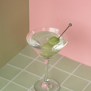 May include: A clear martini in a stemmed glass with two green olives on a pick. The drink sits on a green and white tiled surface, with a pink and tan background.