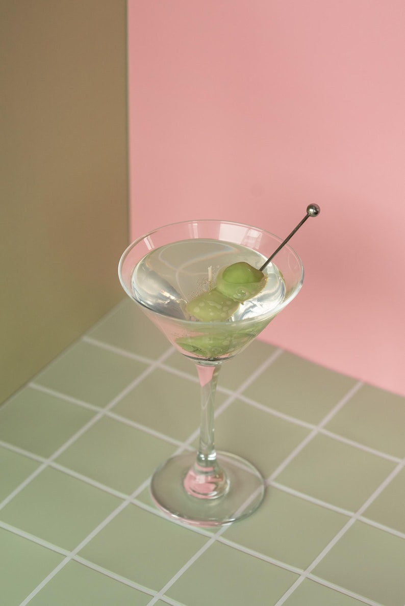 May include: A clear martini in a stemmed glass with two green olives on a pick. The drink sits on a green and white tiled surface, with a pink and tan background.