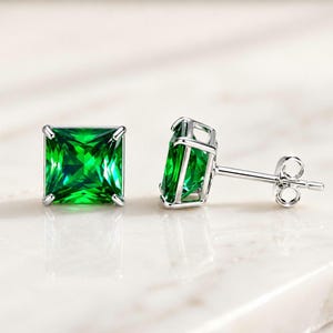 May include: A pair of stud earrings featuring square-cut, vibrant green gemstones set in polished silver-toned metal. One earring is shown from the front, while the other is angled to display the four-prong setting and the post with a butterfly back.